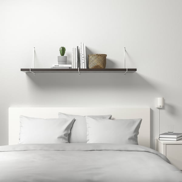 Minimalist white bedroom featuring BERGSHULT wall shelf with books, plant, and basket, above a neatly made bed with white bedding.