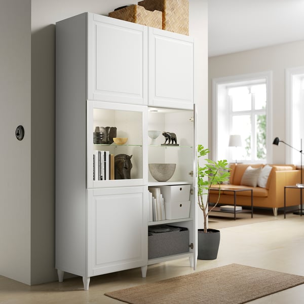 White BESTÅ cabinet with glass doors displays decor and stores items. Adjustable shelves and hinges, soft-close feature shown.