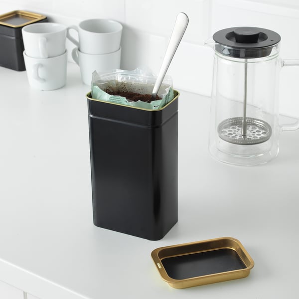 Black metal tin with golden edge stores coffee, protecting it from light and oxygen. Beside it, a glass cafetière and white mugs on counter.