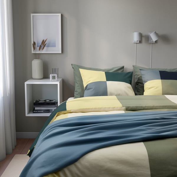 Made bed with BRUNKRISSLA pillows, chequered green, yellow, blue. Blue duvet, grey walls, framed picture, lamp, dried flowers, books, natural light.