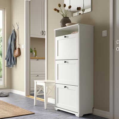 A white shoe cabinet in entryway with adjustable shelves. Nearby are a small stool, jacket, handbag, and vase with flowers.