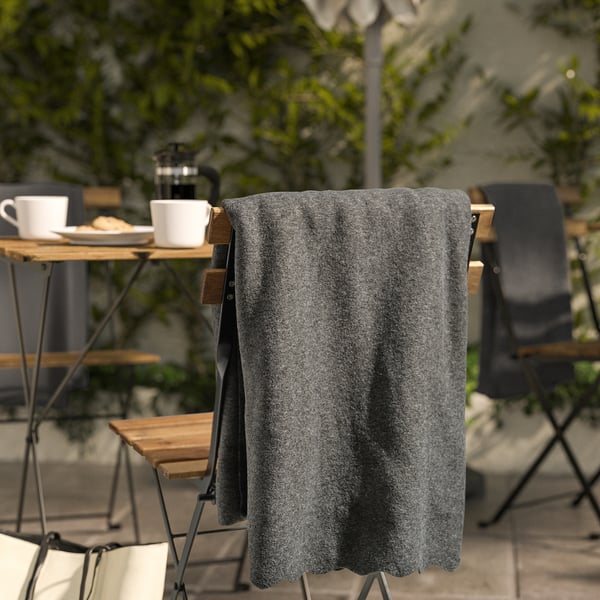 Dark grey DAGGKÅPOR fleece throw draped over chair, larger than cups and plates on wooden table near green plants.