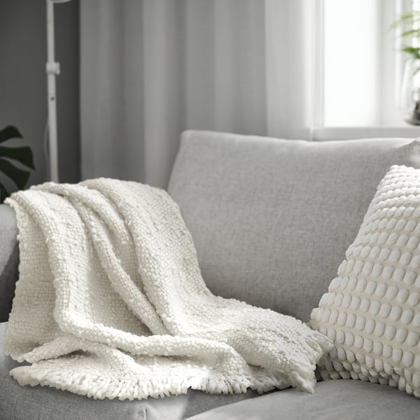 A white DYSTERMAL throw blanket on a grey sofa. The blanket is textured, has fringes, and looks cosy. Its larger than the pillow it covers partially.