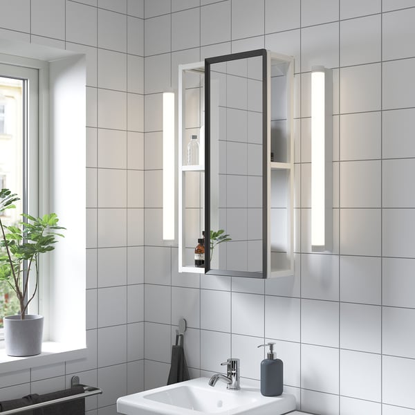Modern bathroom with large ENHET mirror cabinet. White walls, plants, white sink, soap dispenser. Mirror reflects light.