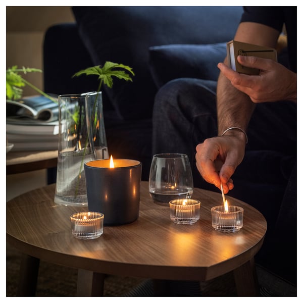 Person lights candle next to lit glass candle, wine glass, and vase with plant on dark wooden table.