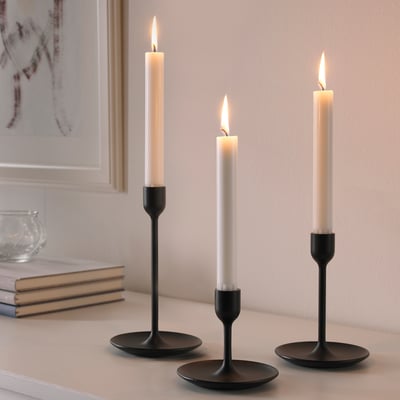 Three black candlesticks of varying heights, each holding a lit white candle, arranged on a table.