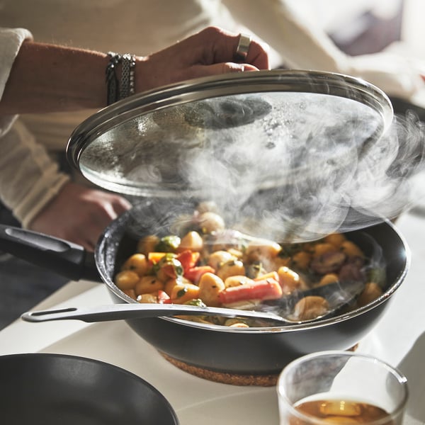 Person lifts lid from sizzling pan, releasing steam. Shows cooking with HEMLAGAD, even heat distribution, steam braising food.