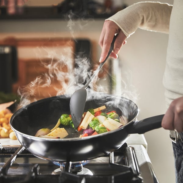 Person cooking vegetables in a non-stick frying pan on a stove, steam rising.