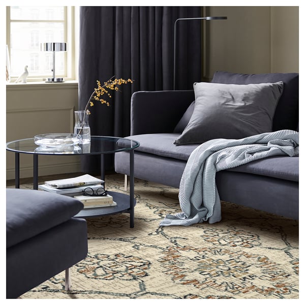 Modern living room with grey HOLSTEBRO rug, blue sofa, glass coffee table, and patterned curtains. Visible decorative vase, books, and blanket.