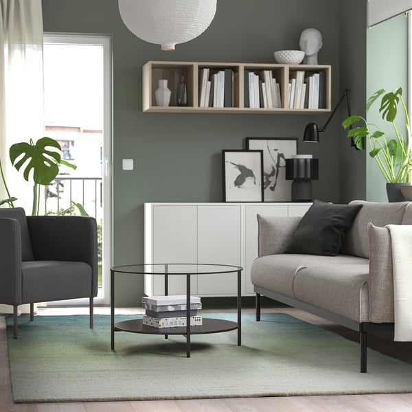 Modern living room with blue-green rug, grey couch, glass table, green walls.