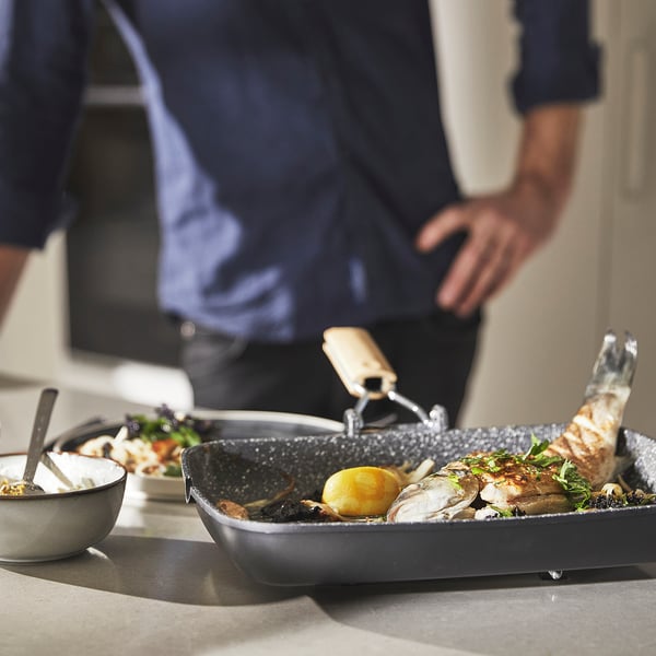 A person is cooking seafood in a non-stick, grey skillet with a lemon and herbs. The pan sits on a countertop, alongside a bowl with suspension sauce.