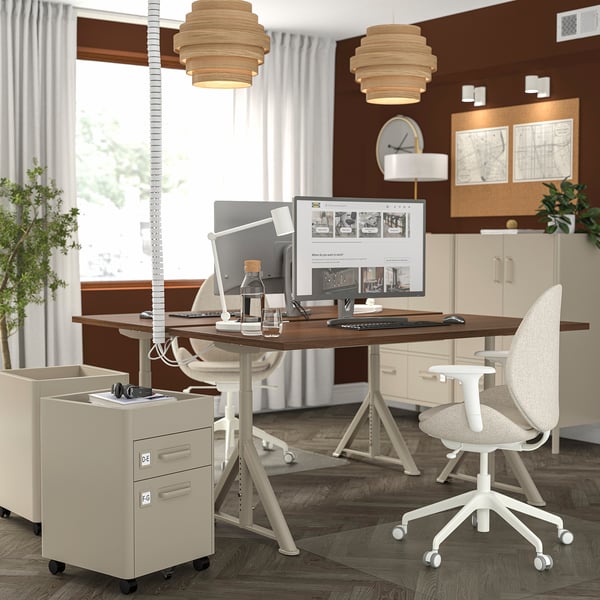 Modern office setup with IDÅSEN desks featuring wooden tops and adjustable white legs. Two computers, chairs, and unique pendant lights.