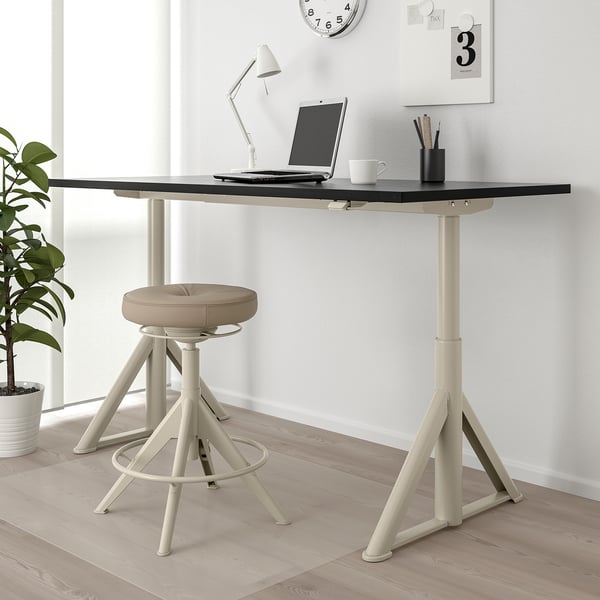 An IDÅSEN beige home office setup featuring a height-adjustable desk with a laptop, stool, and decorations.