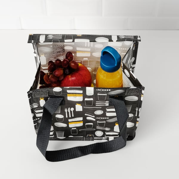 A black ikea bag with kitchen item patterns holds an apple, grapes, and a yellow bottle with a blue cap.