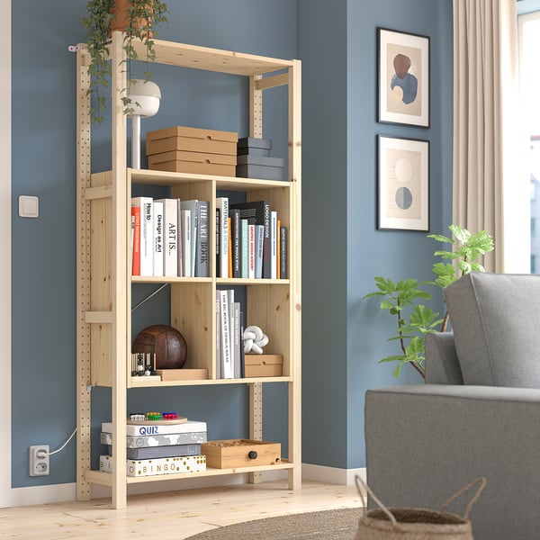 IVAR light pine bookcase stands tall near a blue wall, filled with books and boxes. Near a grey chair, its large and sleek.