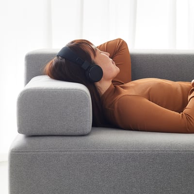 A person relaxes on a grey JÄTTEBO sofa, wearing headphones, with an orange blanket.