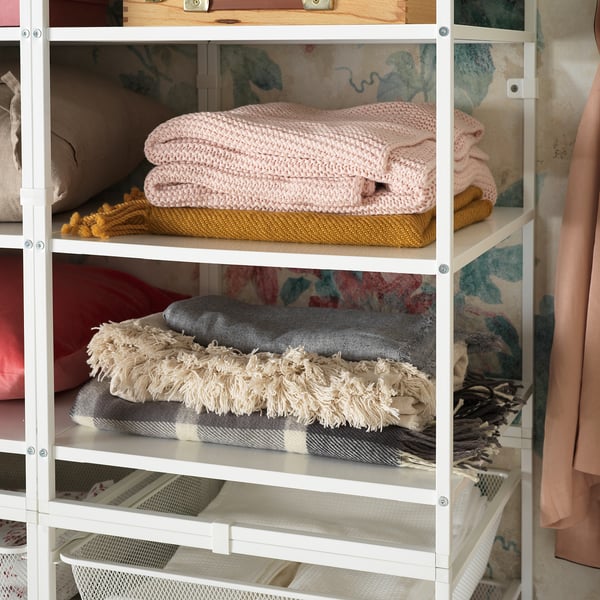 White metal storage shelf holding folded pink, grey, and beige blankets and towels. Neatly organised in individual sections.