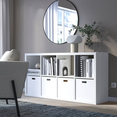 White ikea KALLAX shelf with books, decor, plant, and lamp. Large mirror reflects window. Grey sofa, rug, cozy minimalist space.