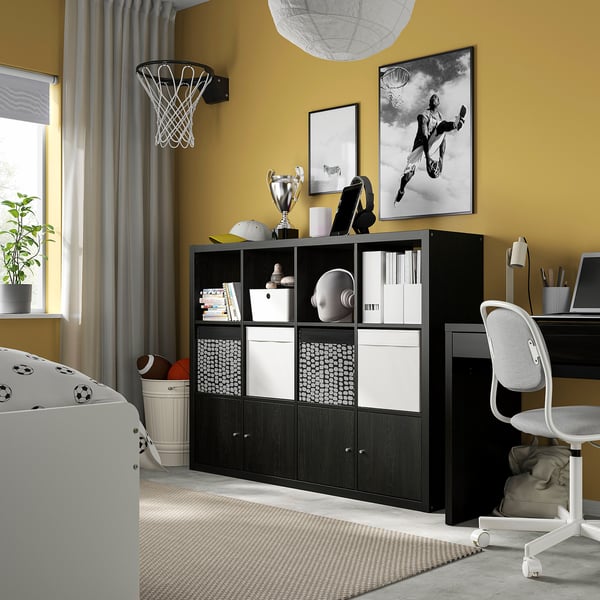 KALLAX black shelf unit beside desk, with white bins for sports gear. Modern design stands out against yellow wall, with basketball hoop above.