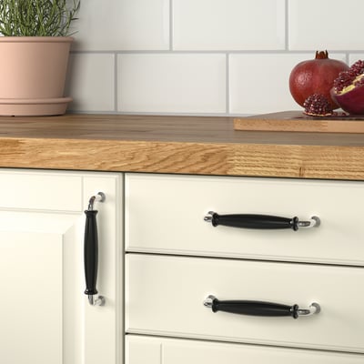 Kitchen drawers with black porcelain knobs, white cabinets, wooden countertop, potted plant, and fresh pomegranates.