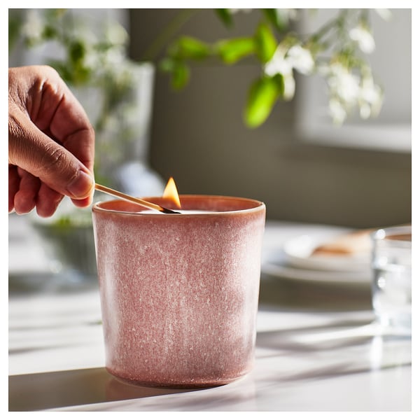 A hand trims a lit wick of a pinkish-brown LUGNARE scented candle in a ceramic jar, creating a romantic atmosphere.