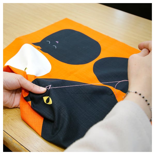 Image shows hands sewing an orange MÄVINN cushion cover with black cat design on fabric.