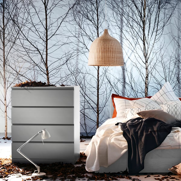 Awesome room with grey MALM dresser, tall wicker lamp, bed with white pillows, and cosy blankets, all surrounded by leafless trees in snow.