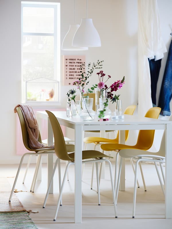 Bright dining room with white table, yellow chairs, colourful flowers, and a hanging white MELODI lamp providing directional light.