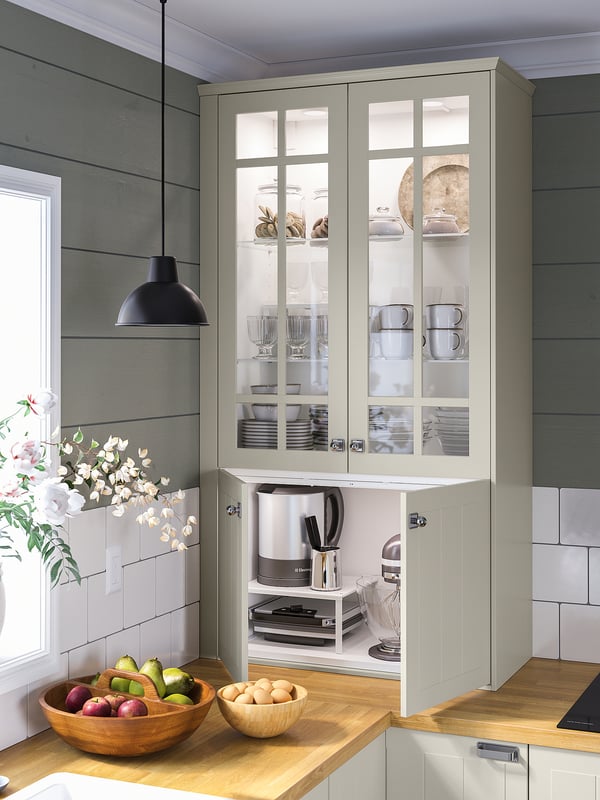 Kitchen with light grey glass-front cabinets, white counter, wooden bowl with fruits, and pendant lamp.