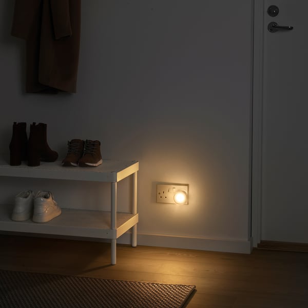 A minimalist entryway with MÖRKRÄDD black lamp on wall beside white shoe rack. Small, discreet light, brightens dark floors.