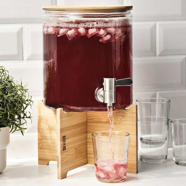 Glass drink dispenser on bamboo stand pours red beverage into glass.