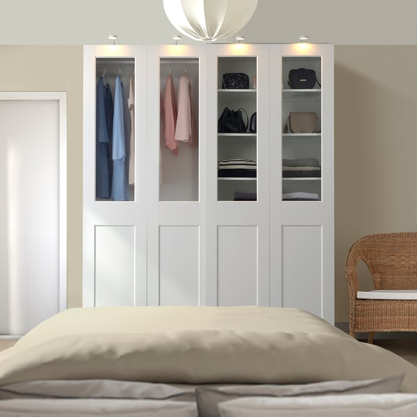 White PAX wardrobe with glass doors, showing clothes and accessories, beside a bed.