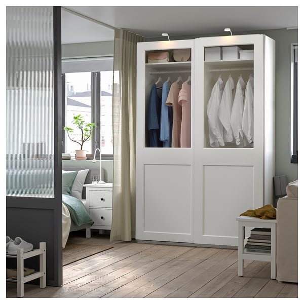Modern white PAX wardrobe with glass doors, displaying clothes. Soft closing, wooden floor setting.