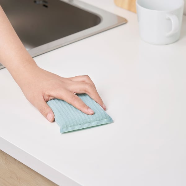 Person cleans counter with light blue PEPPRIG scrubber. Sink and cup visible in background.