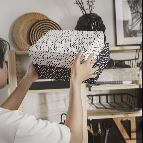 Person placing dotted storage box on shelf near workspace. The box helps organise papers and desk accessories efficiently.