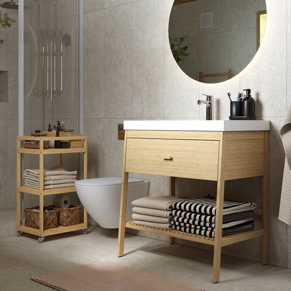 Modern bathroom with RÅGRUND bamboo vanity, white sink, round mirror, storage cart, and striped towels.