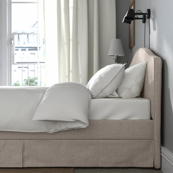 Elegant bed with grey cover, two white pillows, and a table lamp. Wall-mounted light and big window, modern room.