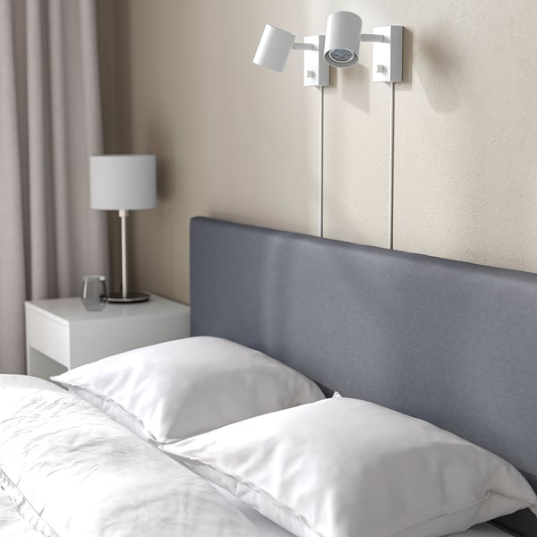 Minimalist bed with blue headboard, wall-mounted light, white linens, and bedside table with lamp.