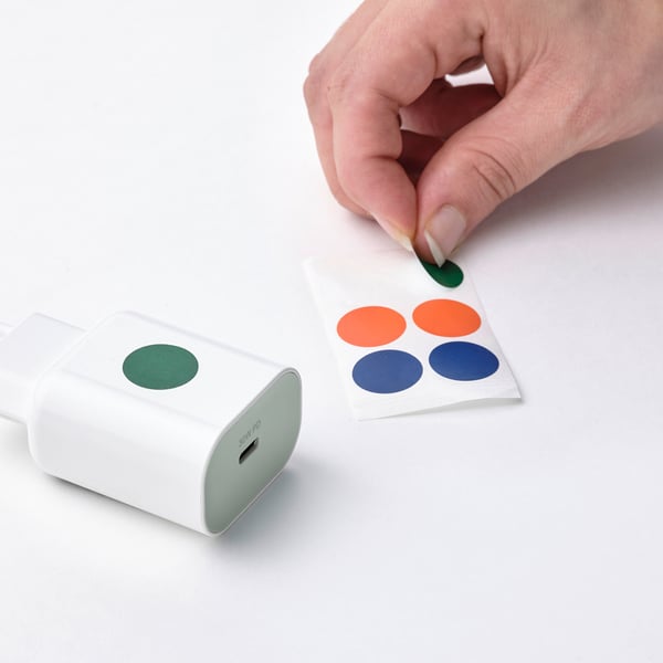 Image shows a hand personalising a white usb-c charger with green sticker, beside colourful dot stickers.