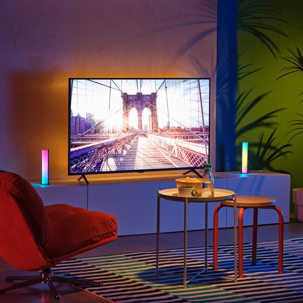 Modern tv in colourful living room with brooklyn bridge on screen. Red chair, tables, rug, plants, colourful lights.