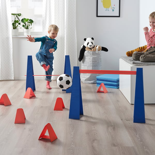 Kids playing with SKRATTMÅS obstacle course, jumping, crawling, and balancing with cones and hurdles in a living room.