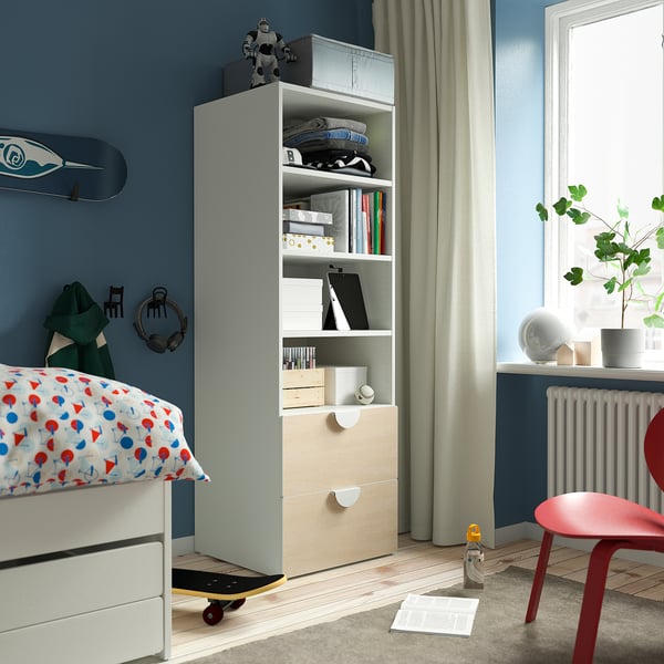 Modern blue bedroom with white bookcase holding items, red chair, colourful bed pillows.