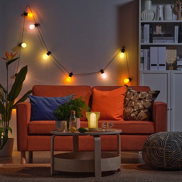A cosy living room with orange couches, string lights, and a coffee table holding drinks and a vase.