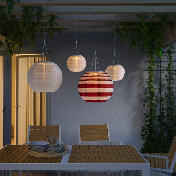 Four SOLVINDEN lanterns in red, white, & other colours illuminate an outdoor dining area under a pergola.