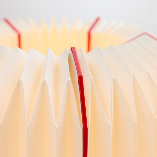 Modern table lamp with pleated, cream-colored shade, accented by red lines, casting warm, inviting light.