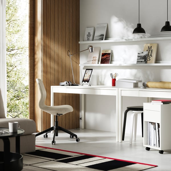 Modern home office with white desk, chair, and shelves. Drawer and shelves hold books and decor. Natural light and a red/black rug.