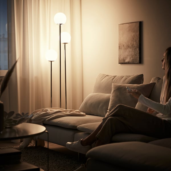 A cosy living room with a person reading on a beige couch under three globe floor lamps, illuminated curtains, and a framed wall art.