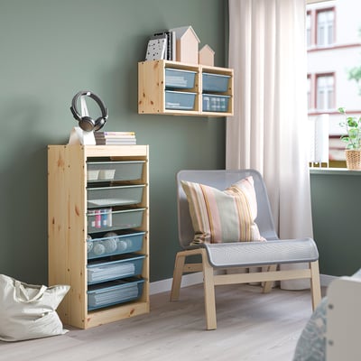 Wooden TROFAST unit with mesh drawers, holding books and headphones, paired with a cushioned chair and wall shelf for organisation.