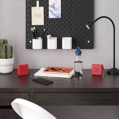 Modern workspace with black desk, VAPPEBY speaker, books, smartphone, water bottle, pegboard with supplies, cactus, and lamp.