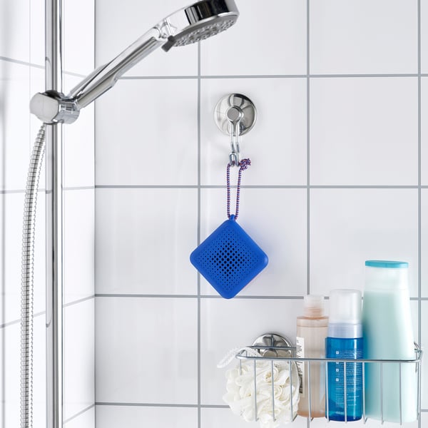 A blue waterproof bluetooth speaker hangs in a shower. Nearby, toiletries like shampoo and body wash are on a chrome wire shelf.